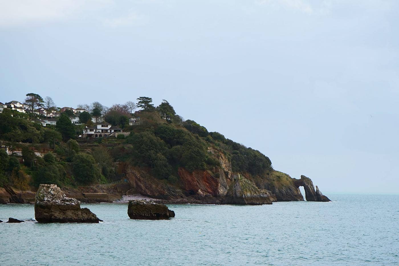 Homes on the wooded hillside above the sea at Torquay, with a rock arch in the water Homes on the wooded hillside above the sea at Torquay, with a rock arch in the water